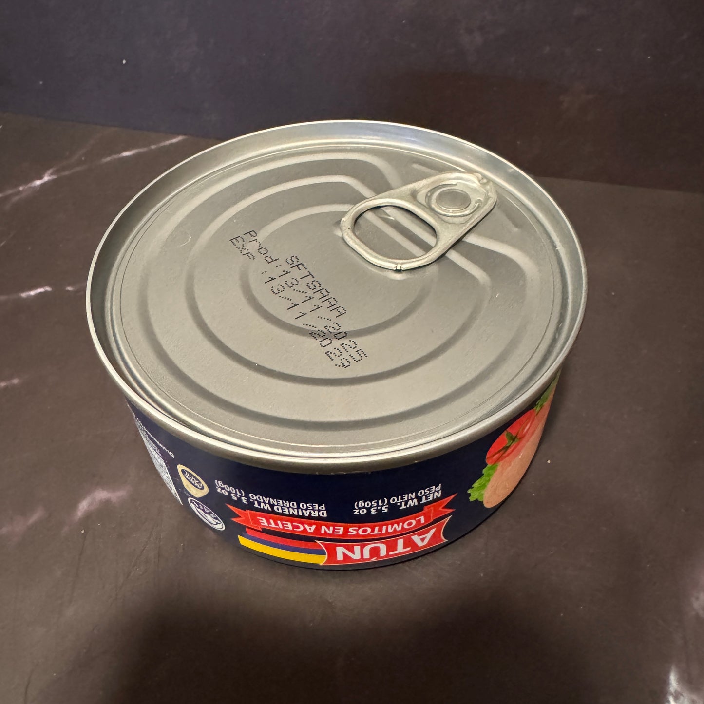 A can of Atun Lomitos en Aceite (fish and chips in oil) on a dark brown countertop. The can features a dark blue label with red and yellow accents, and a silver metal lid with a pull-tab design.