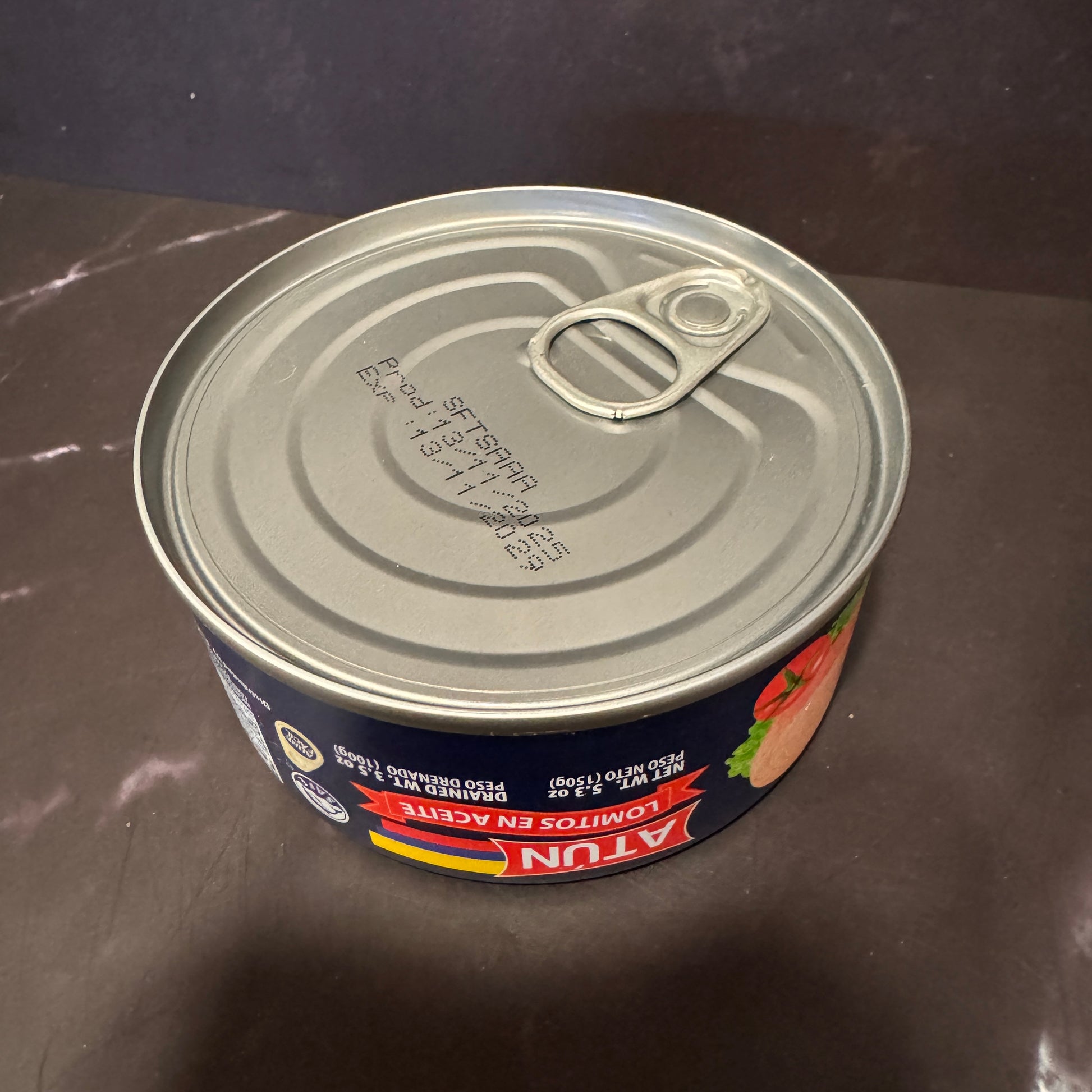 A can of Atun Lomitos en Aceite (fish and chips in oil) on a dark brown countertop. The can features a dark blue label with red and yellow accents, and a silver metal lid with a pull-tab design.