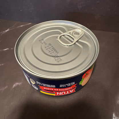 A can of Atun Lomitos en Aceite (fish and chips in oil) on a dark brown countertop. The can features a dark blue label with red and yellow accents, and a silver metal lid with a pull-tab design.